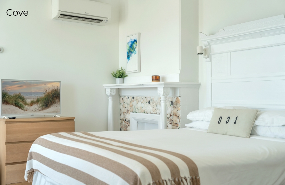 A minimalist, beach-themed bedroom with a white bed, a tan and white striped throw, a wooden dresser with a flat-screen TV, and a white mantle decorated with sea stones. A wall-mounted air conditioning unit is positioned above the dresser.