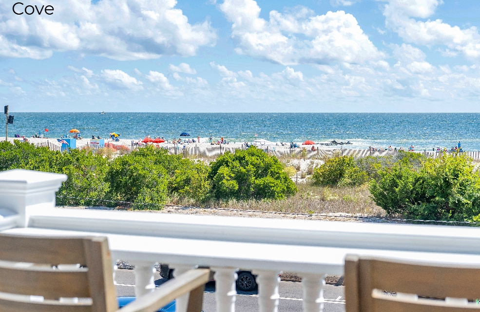 A view from a white-railed balcony looking out over a sandy beach populated with colorful umbrellas and people, with the blue ocean and a cloudy sky in the background.