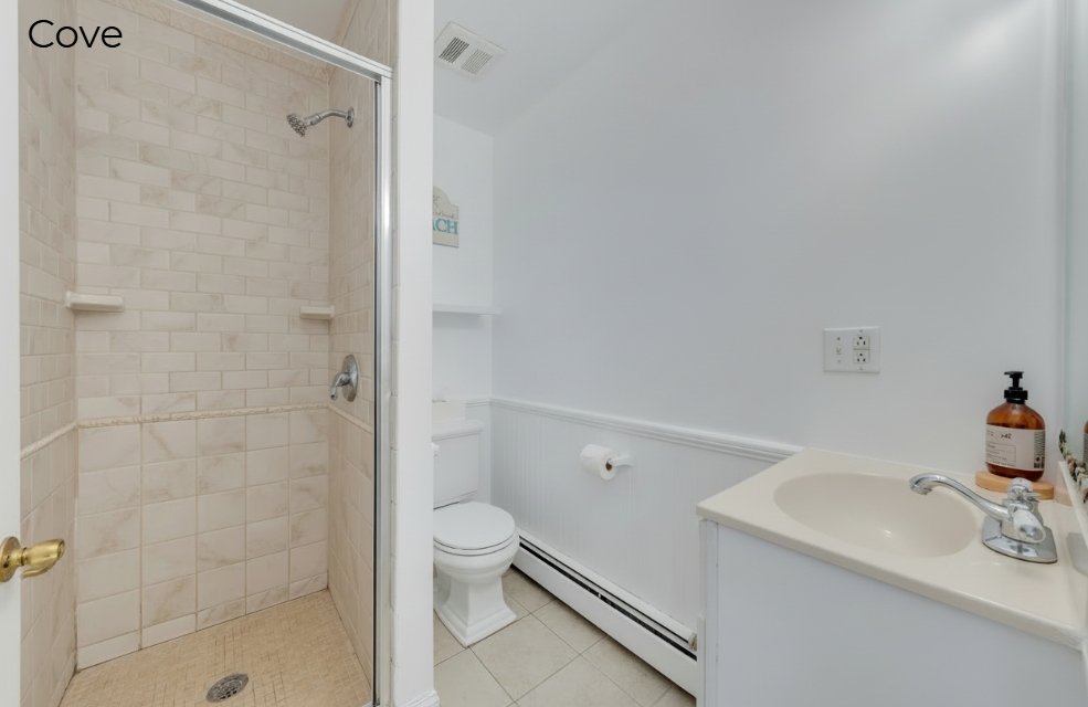 A simple, white-walled bathroom featuring a glass-door walk-in shower with tan subway tiling, a white toilet, and a small vanity with a cream-colored countertop.