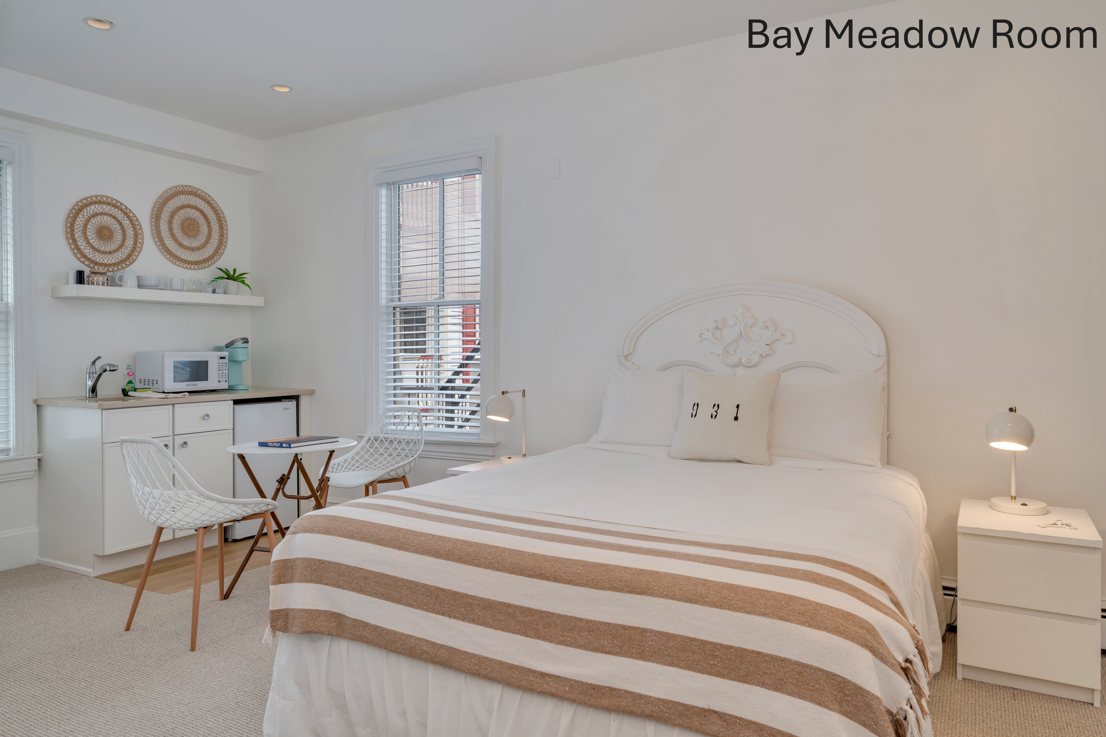 A spacious white bedroom featuring a white-skirted bed with a tan and white striped throw, a brown armchair, and a decorative fireplace with a mosaic tile surround. A small kitchenette area and a window with blinds are visible in the background.