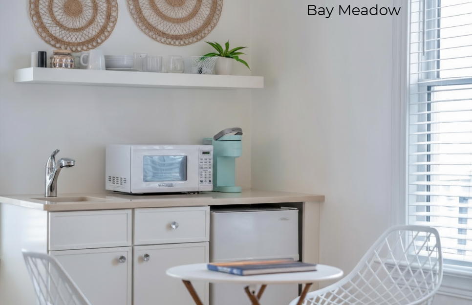 A white-walled kitchenette featuring a white microwave, a light-blue coffee maker, and a mini-fridge under a tan countertop. Above, a white floating shelf holds decorative items, and a small round table with two white mesh chairs is positioned in the foreground.