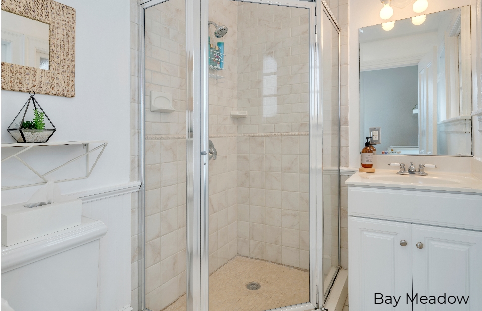 A minimalist bathroom featuring a white vanity and toilet, a mirror with a light fixture, and a glass-enclosed corner shower with beige subway tiles.