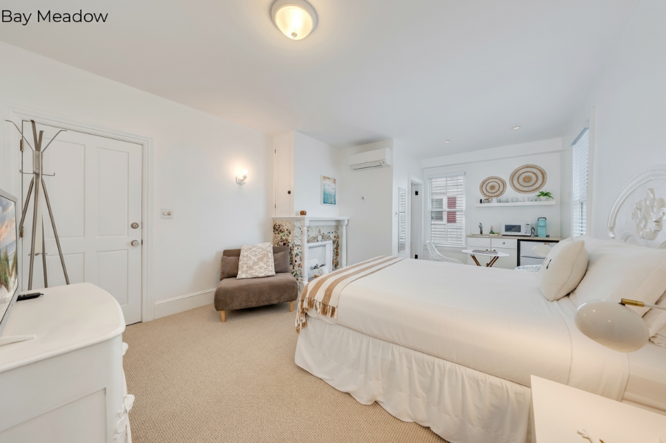 A white-walled bedroom featuring a white-skirted bed with a striped throw, a brown armchair, and a decorative tiled mantle. In the background, a small kitchenette area with wall decor is visible next to a window.