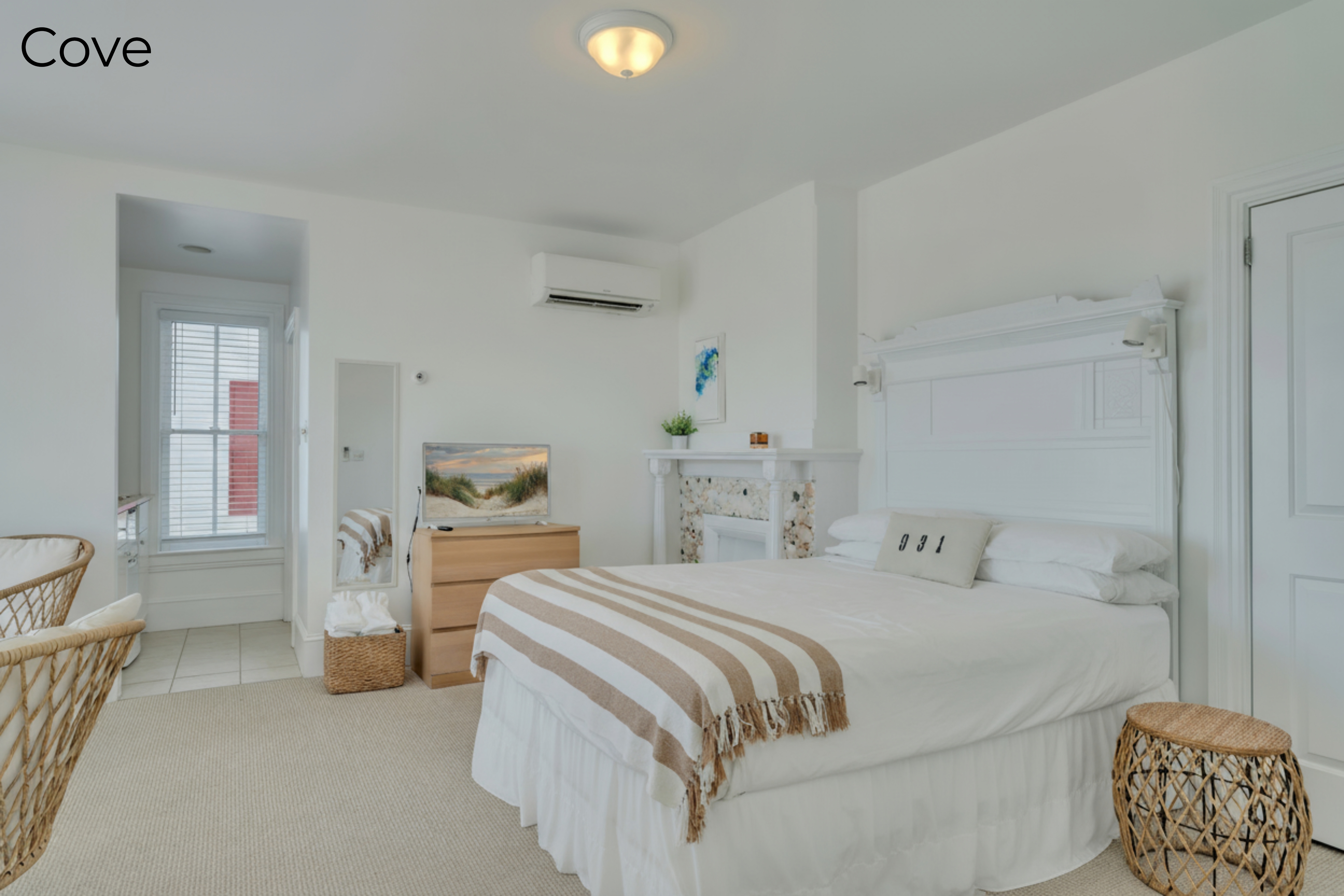 A bedroom featuring a bed with a tan and white striped throw, a wooden dresser with a flat-screen TV, and a decorative fireplace mantel with a mosaic tile surround. To the left, a partial view of woven wicker chairs and an open doorway leading to a brightly lit kitchenette area are visible.