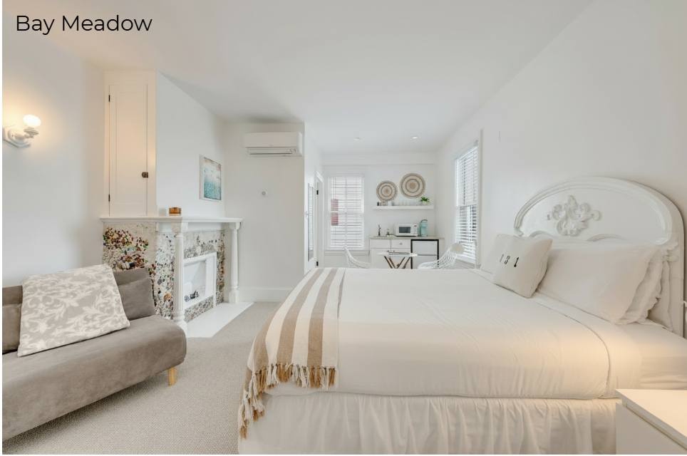 A spacious white-walled bedroom labeled "Cove" featuring a white-skirted bed with a tan and white striped throw. The room includes a light-colored wooden dresser with a flat-screen TV, a decorative mantel with a mosaic tile surround, and a wall-mounted air conditioning unit. To the left, woven wicker chairs and an open doorway leading to a brightly lit kitchenette area are visible.