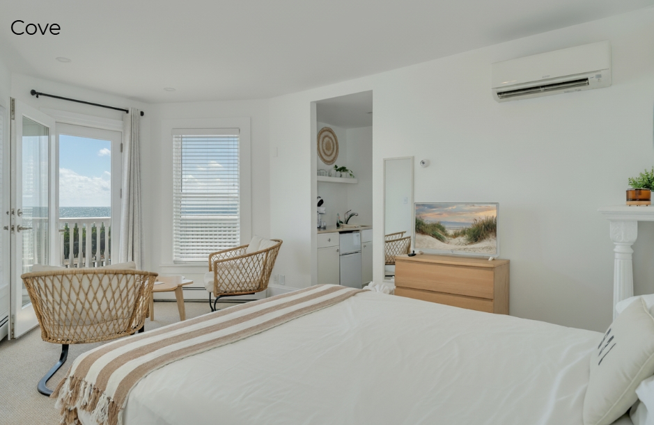 A bright, minimalist bedroom featuring a white bed with a tan and white striped throw, a wooden dresser with a flat-screen TV, and a decorative fireplace mantel with a mosaic tile surround. To the left, two woven wicker chairs face a window and glass door that open onto a balcony overlooking the ocean.