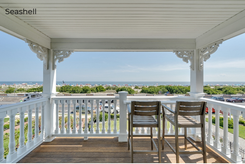A view from a covered wooden porch labeled "Seashell," featuring a white railing with a built-in bar top and two wooden bar-style chairs. The balcony overlooks a green lawn and a beach dotted with umbrellas, with the blue ocean and a clear sky visible in the distance under decorative white architectural brackets.