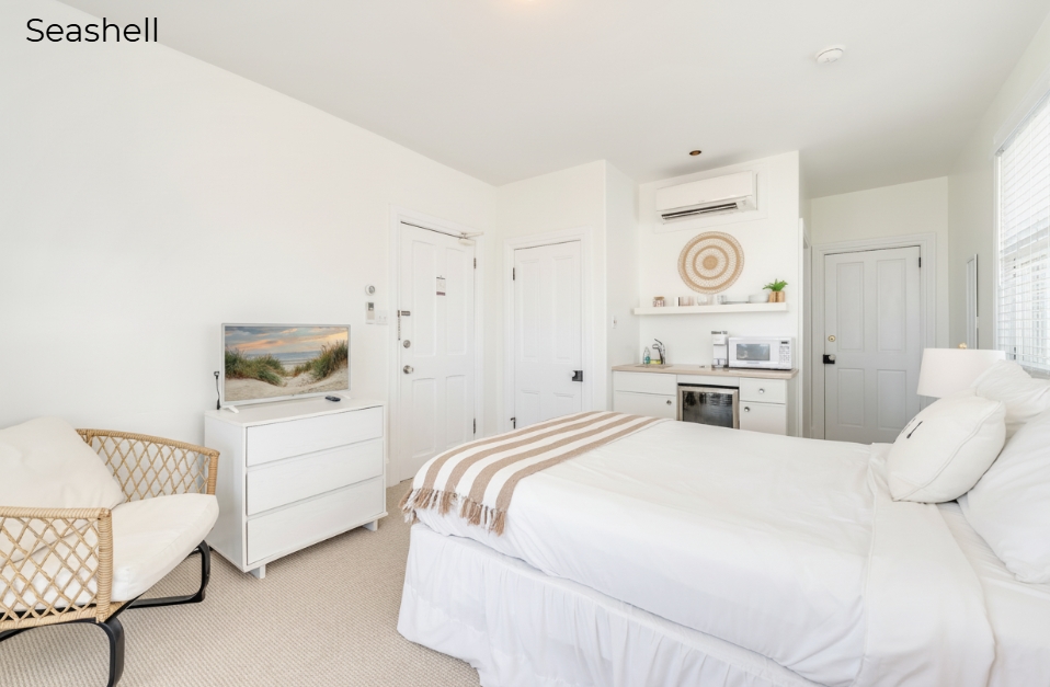 An interior view of the minimalist Seashell bedroom featuring a white bed with a tan and white striped throw in the foreground. To the left, a woven wicker chair sits beside a white three-drawer dresser topped with a flat-screen TV, while the background shows an open-plan layout leading to a small kitchenette with a mini-fridge and white cabinetry.