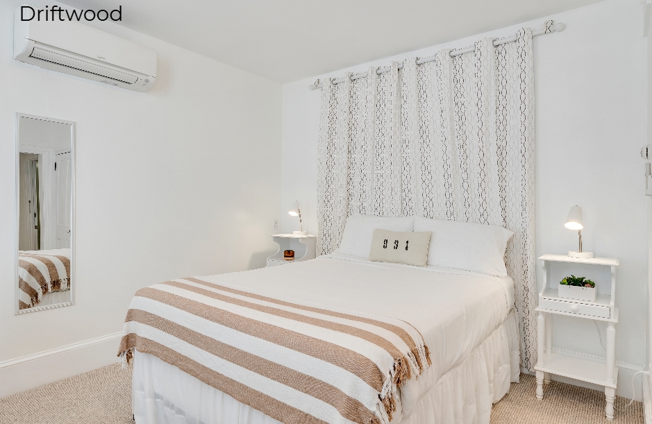 A bright, minimalist bedroom in the Driftwood suite featuring a white bed with a tan and white striped throw. The bed is positioned against a wall with floor-to-ceiling patterned curtains, flanked by a small white nightstand with a lamp, while a tall mirror and wall-mounted air conditioning unit are visible on the left.