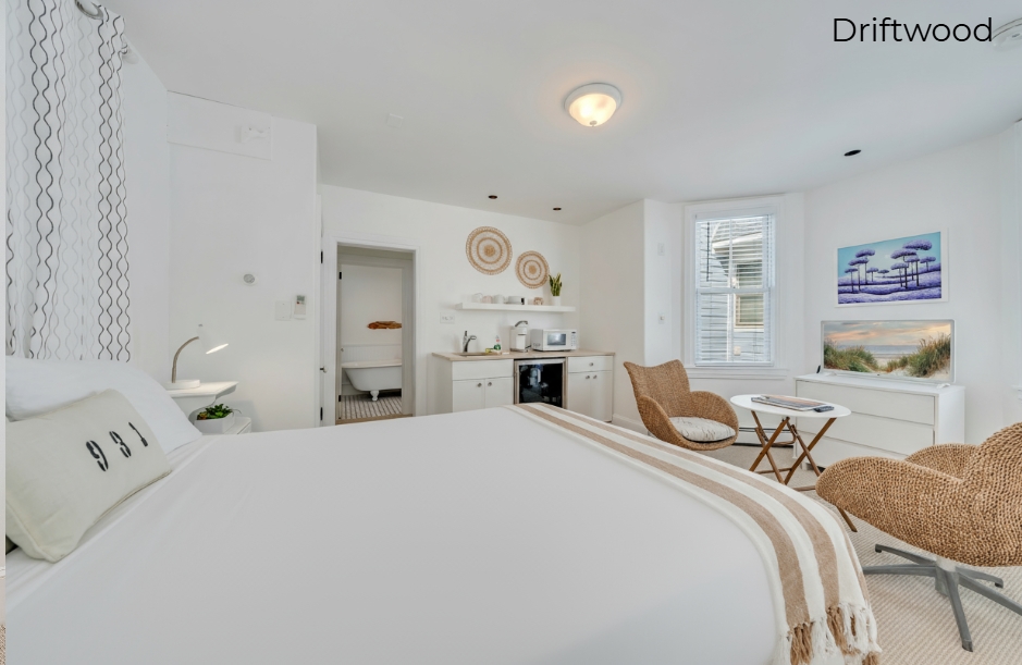 A minimalist bathroom in the Driftwood suite featuring a white vanity with a tan countertop, a white toilet, and a glass-enclosed walk-in shower with beige subway tiling and a black and white tiled floor.