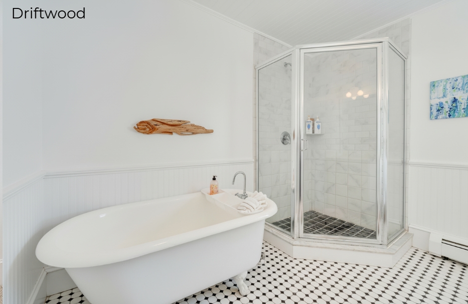 A spacious white-walled bathroom in the Driftwood suite featuring a white clawfoot bathtub and a glass-enclosed corner shower with marble-patterned walls and a black-and-white tiled floor. Above the tub, a piece of natural driftwood is mounted on the wall as decor.