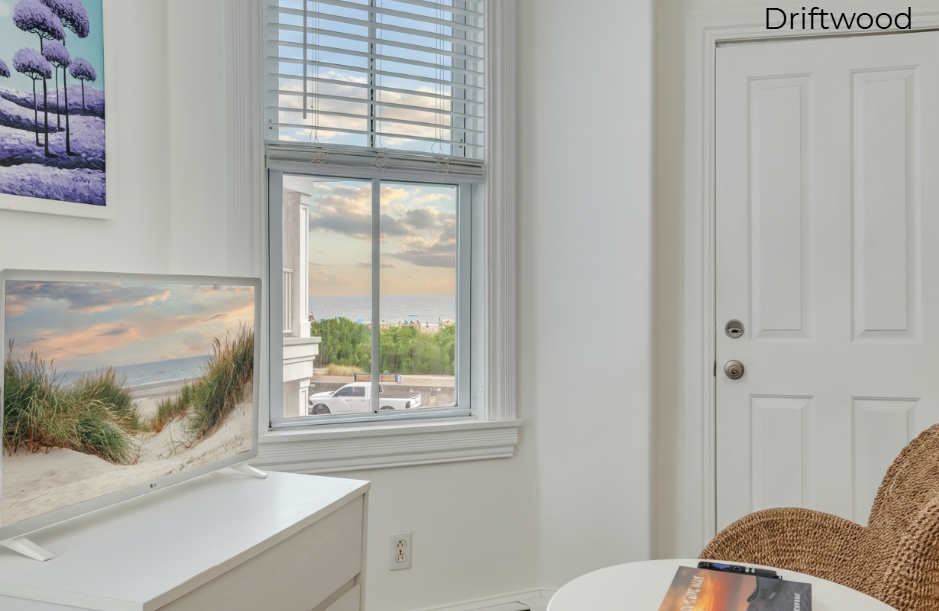 A close-up view of the "Driftwood" suite, showing a white three-drawer dresser with a flat-screen TV in the foreground. A window with white blinds reveals a glimpse of the beach and ocean at sunset, and a white door and the edge of a woven wicker chair are visible on the right.