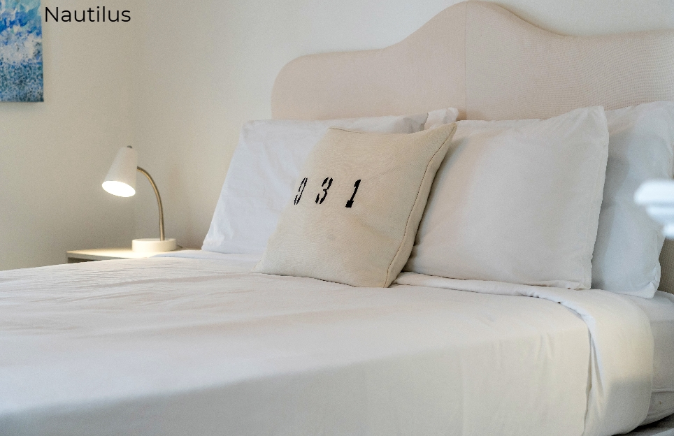 A close-up view of a bed with white linens and a cream-colored arched headboard. A decorative beige pillow featuring the numbers "931" sits in front of standard white pillows, and a small white bedside lamp is illuminated in the background.