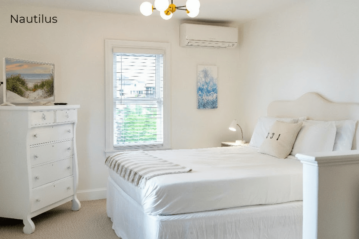 A brightly lit bedroom area within a studio apartment featuring a large bed with white linens and a cream arched headboard. To the left, there is a white dresser topped with beach-themed artwork, a window with white blinds, and a modern gold and glass chandelier hanging from the ceiling.