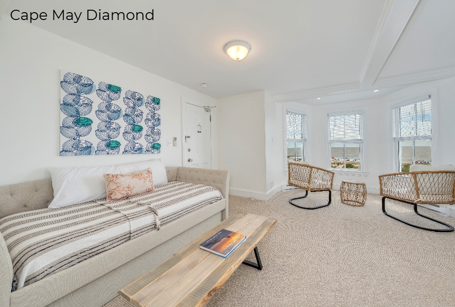 A spacious living area with white walls and light beige carpeting. The room features a neutral-toned daybed topped with a striped blanket and a small patterned pillow, set against a wall with a blue and white botanical art print. Two modern wicker armchairs are positioned near a large bay window that offers a view of the outdoors.