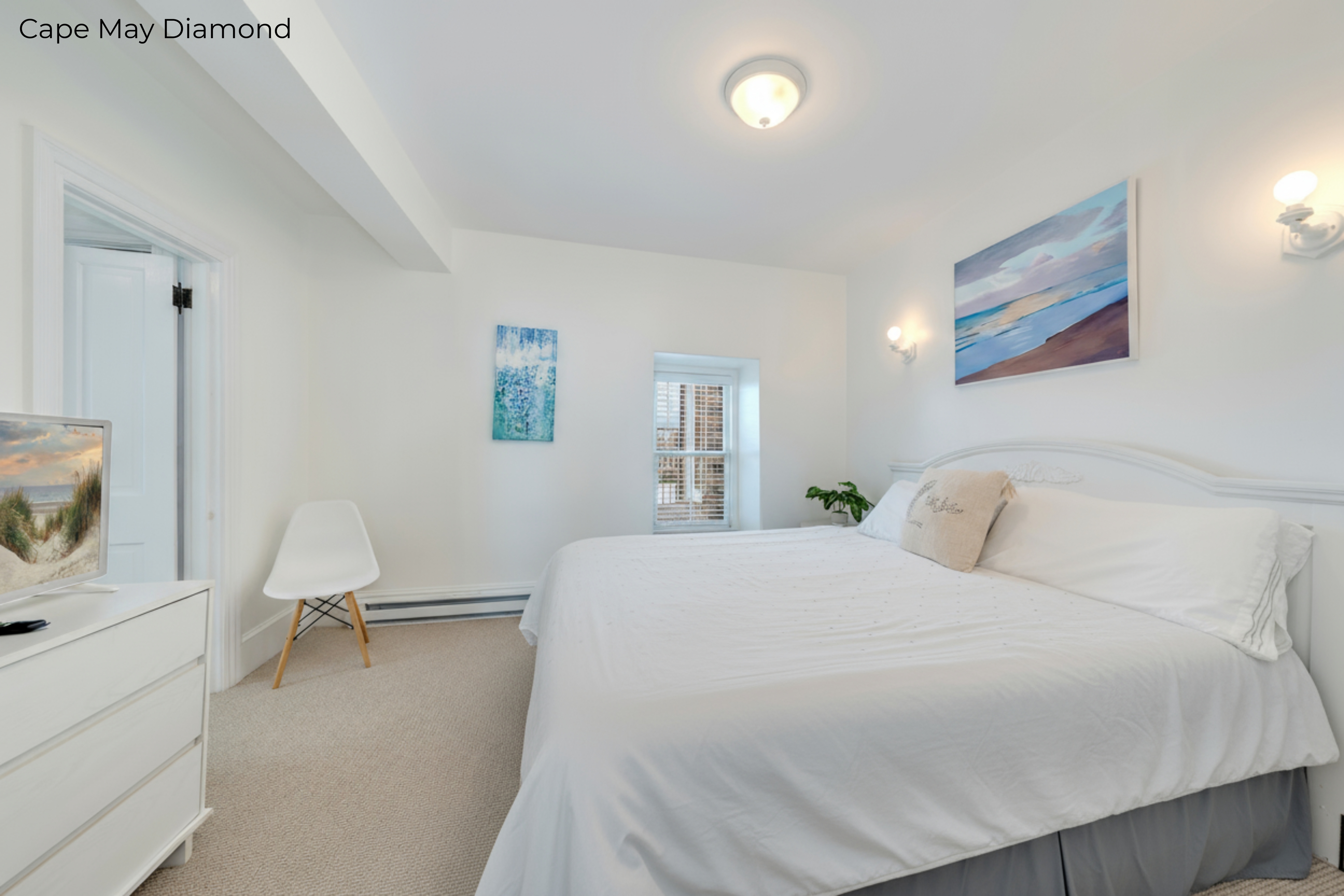 A bright bedroom with white walls and beige carpeting features a bed dressed in crisp white linens and a grey bedskirt. Above the arched white headboard hangs a horizontal painting of a beach sunset, flanked by two glowing wall sconces. To the left, a white dresser and a modern white chair are visible near an open doorway.