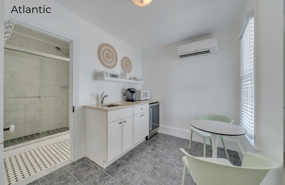 A brightly lit kitchenette featuring white cabinetry and light wood countertops, with a coffee maker and microwave. The space includes a small, round white dining table with two light green chairs, set against a window with white blinds and grey-tiled flooring. To the left, an open doorway reveals a modern bathroom with a glass-enclosed walk-in shower.