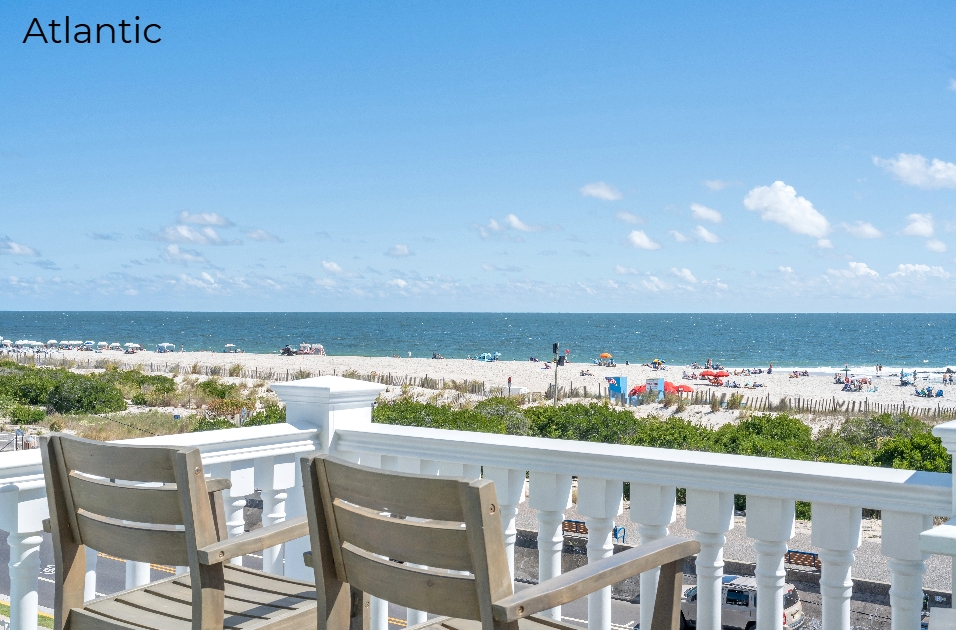 A wide-angle view from a balcony with two wooden chairs overlooking a bright, sunny beach. Beyond the white railing, the scene features sandy dunes with green shrubs, a populated beach with colorful umbrellas, and a calm blue ocean under a clear sky with scattered white clouds.