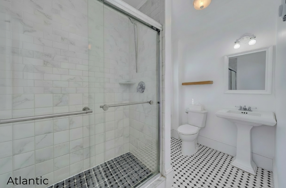 A bright, modern bathroom featuring black and white patterned floor tiles and white walls. On the left, a large walk-in shower is enclosed by sliding glass doors and lined with white marble-style tiles. The right side of the room includes a white pedestal sink with a framed mirror above it, a white toilet, and a simple wooden floating shelf.