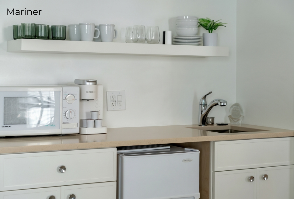 A white kitchenette with light wood countertops featuring a small sink, a coffee maker, and a white microwave. Above the counter, a white floating shelf holds glassware and small plants, while a compact white refrigerator is built into the lower cabinetry.