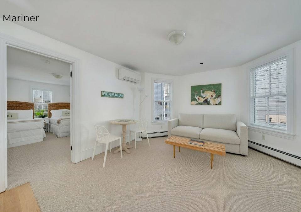 A bright living area featuring a light beige sofa with matching pillows, a wooden coffee table, and beige carpeting. The room has white walls, two windows with blinds, and an open doorway leading to a bedroom with two beds. A small round dining table with two white geometric chairs sits against the wall beneath a green "MERMAIDS" sign.