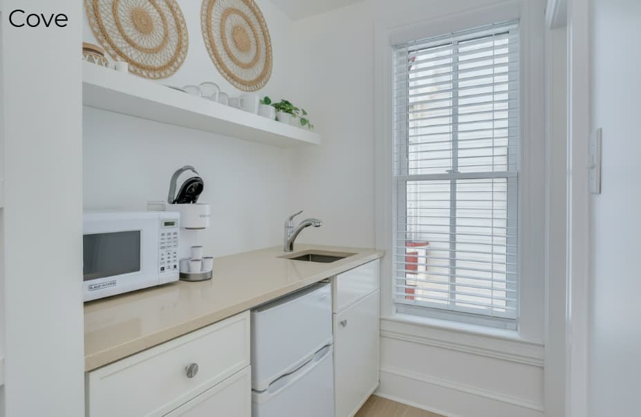 A compact white kitchenette featuring a tan countertop with a small sink, a white microwave, a coffee maker, and a mini-fridge, situated next to a window with white blinds.