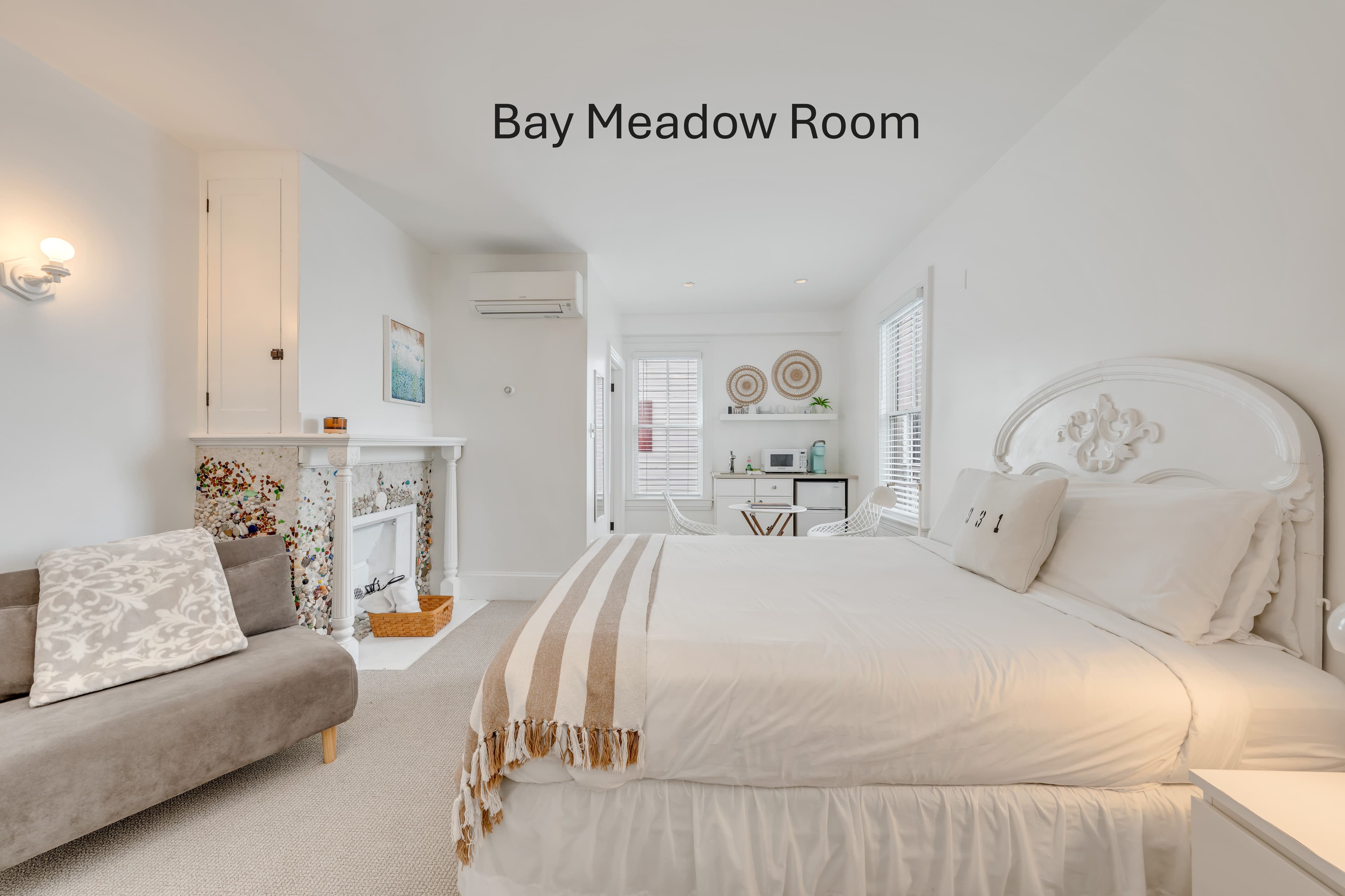 A spacious white bedroom with a white-skirted bed, a brown armchair, and a small kitchenette area in the background under a ceiling light.