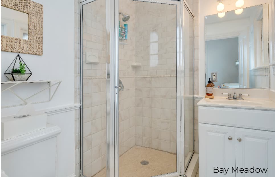 A wide-angle view of a minimalist, white-walled bathroom in the Bay Meadow suite featuring a white vanity with a silver faucet, a large mirror with warm light fixtures, and a glass-enclosed walk-in shower with beige subway tiling. To the left, a white toilet is visible beneath a small decorative shelf holding a plant.
