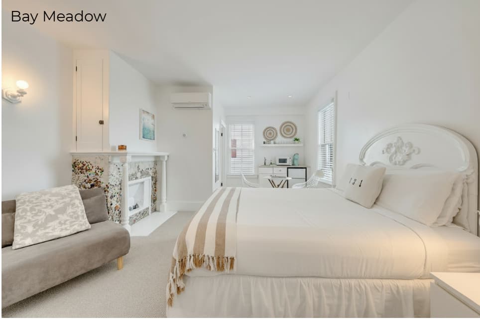 A spacious white-walled bedroom labeled "Cove" featuring a white-skirted bed with a tan and white striped throw. The room includes a light-colored wooden dresser with a flat-screen TV, a decorative mantel with a mosaic tile surround, and a wall-mounted air conditioning unit. To the left, woven wicker chairs and an open doorway leading to a brightly lit kitchenette area are visible.