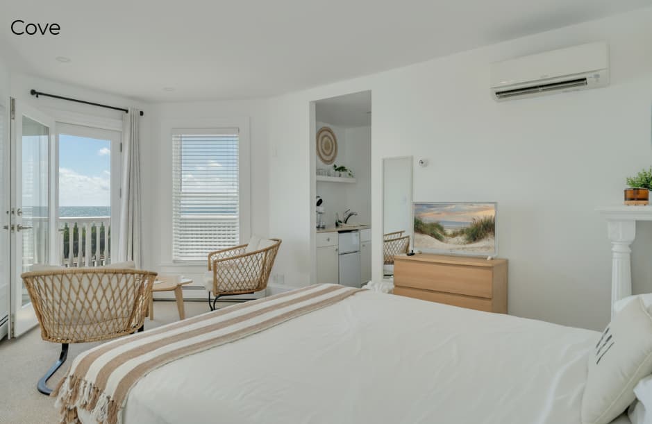A bright, minimalist bedroom featuring a white bed with a tan and white striped throw, a wooden dresser with a flat-screen TV, and a decorative fireplace mantel with a mosaic tile surround. To the left, two woven wicker chairs face a window and glass door that open onto a balcony overlooking the ocean.