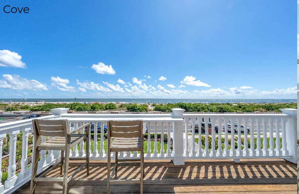 A high-angle view from a white-railed balcony labeled "Cove," featuring two wooden bar-style chairs pulled up to a railing overlooking a green lawn and the beach. In the background, the blue ocean meets a clear blue sky with scattered white clouds.