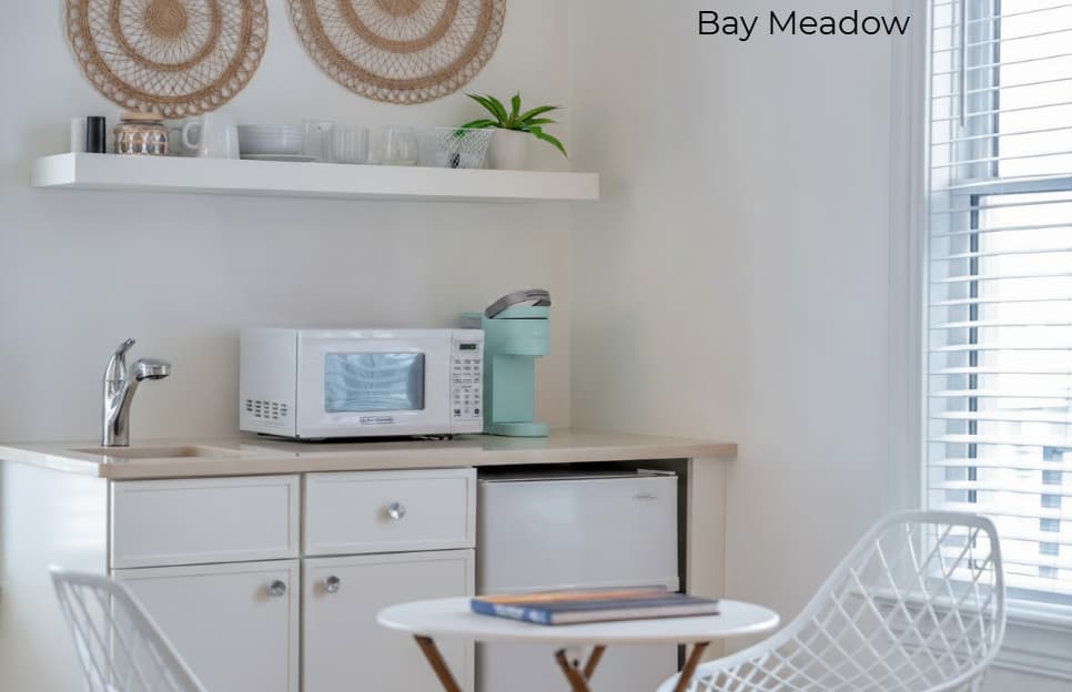 A white-walled kitchenette in the "Bay Meadow" suite featuring a white microwave, a light-blue coffee maker, and a mini-fridge beneath a tan countertop. Above the counter, a white floating shelf displays decorative items, and in the foreground, a small round table is paired with two white mesh chairs.