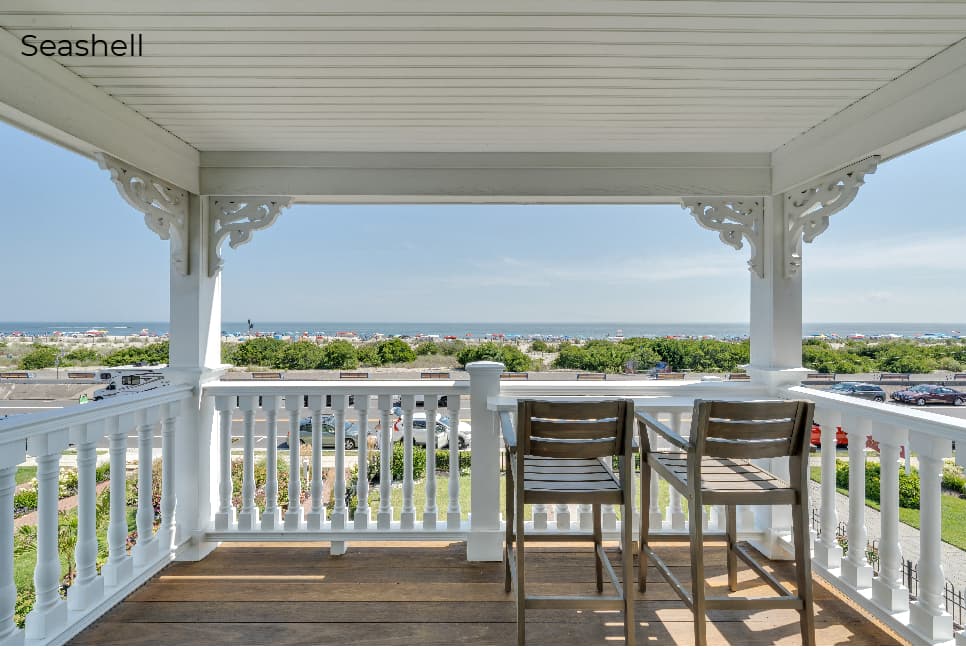A view from a covered wooden porch labeled "Seashell," featuring a white railing with a built-in bar top and two wooden bar-style chairs. The balcony overlooks a green lawn and a beach dotted with umbrellas, with the blue ocean and a clear sky visible in the distance under decorative white architectural brackets.