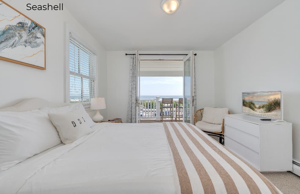 A bright bedroom in the "Seashell" suite featuring a white bed with a striped throw, a white dresser with a flat-screen TV, and an open glass door leading to a balcony with an ocean view.