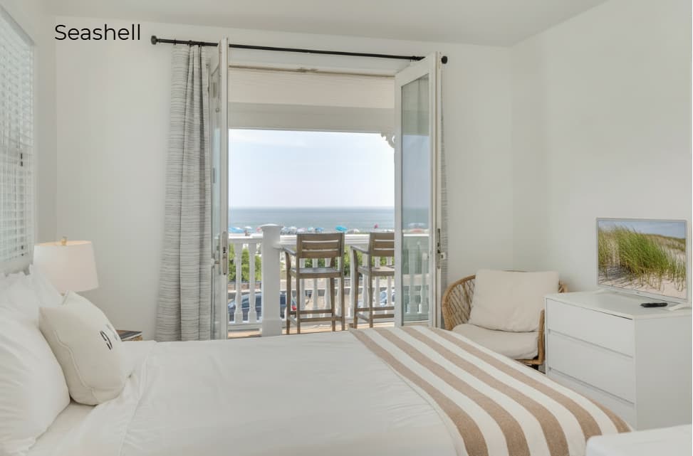 A bright, minimalist bedroom in the Seashell suite featuring a white bed with a tan and white striped throw. An open glass door leads to a private balcony with two wooden bar chairs, offering a direct view of the ocean and the beach beyond.