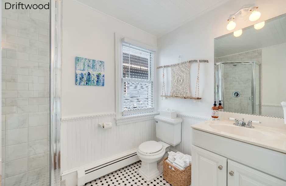 A bright, minimalist bathroom in the Driftwood suite featuring a white vanity with a tan countertop, a white toilet, and a glass-enclosed walk-in shower with marble-patterned tiles and a black and white tiled floor. A window with white blinds is positioned next to the shower, and a large mirror with a warm light fixture hangs above the sink.