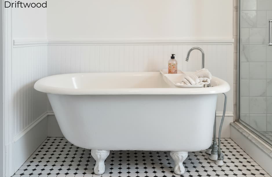 A white-walled bathroom in the Driftwood suite featuring a white clawfoot bathtub and a glass-enclosed corner shower with marble-patterned walls and a black and white tiled floor. Above the tub, a piece of natural driftwood is mounted on the wall as a decorative accent.
