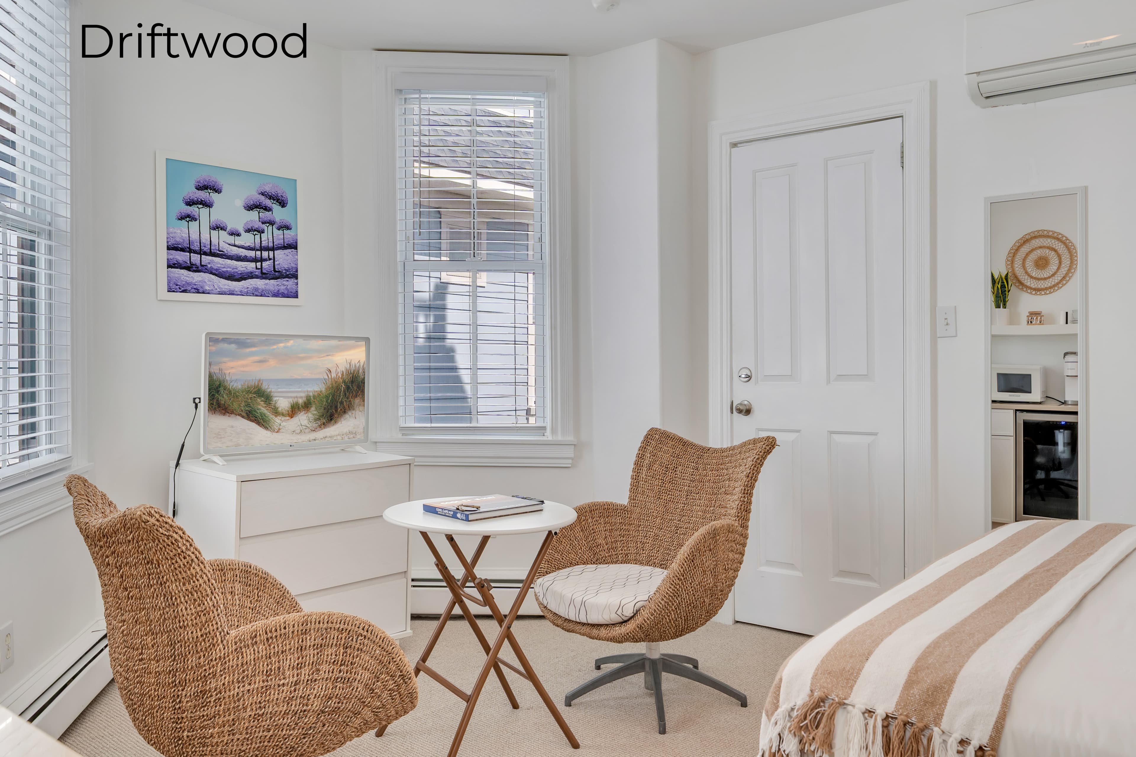 A bright, minimalist bedroom in the Driftwood suite featuring a white bed with a tan and white striped throw. In the foreground, two woven wicker armchairs are positioned around a small white table, and a white dresser with a flat-screen TV sits against the wall near a window. In the background, an open doorway reveals a small kitchenette area with a mini-fridge and wall decor.