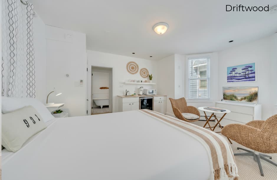 A minimalist bathroom in the Driftwood suite featuring a white vanity with a tan countertop, a white toilet, and a glass-enclosed walk-in shower with beige subway tiling and a black and white tiled floor.