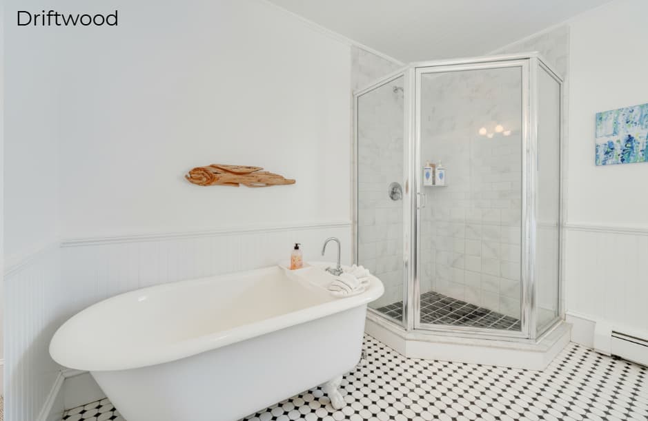 A spacious white-walled bathroom in the Driftwood suite featuring a white clawfoot bathtub and a glass-enclosed corner shower with marble-patterned walls and a black-and-white tiled floor. Above the tub, a piece of natural driftwood is mounted on the wall as decor.