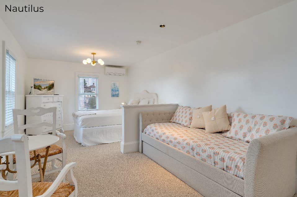 A spacious studio apartment with white walls and beige carpeting. In the foreground, a daybed with patterned orange-and-white pillows is separated from a main bed by a low partial wall. A small white dining set is visible on the left, and a modern globe chandelier hangs from the ceiling.