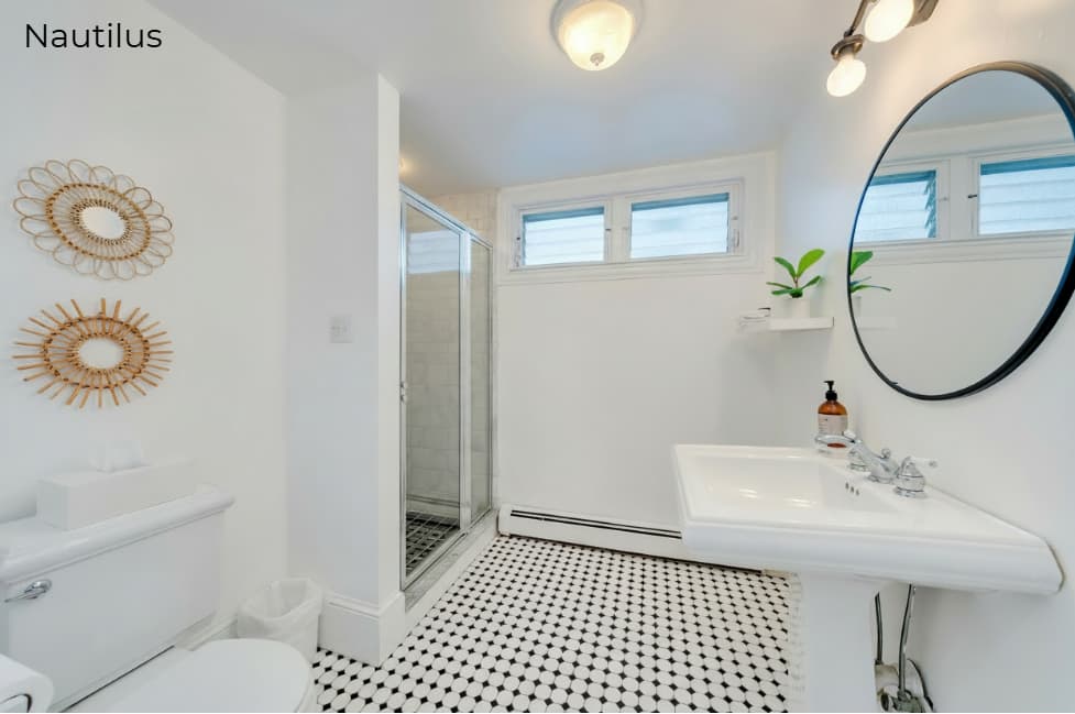 A brightly lit, modern bathroom with black and white patterned tile flooring. It features a white pedestal sink with a round black-framed mirror, a white toilet, and a glass-enclosed walk-in shower. Two circular woven wall hangings and a small potted plant add decorative touches to the clean, white space.
