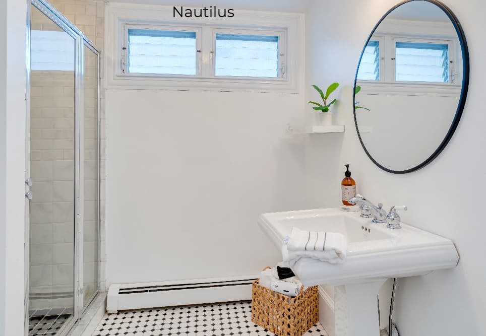 A brightly lit, modern bathroom featuring a white pedestal sink with a round, black-framed mirror above it. The space includes a glass-enclosed walk-in shower on the left, a small window with horizontal blinds, and a wicker basket on the black-and-white patterned tile floor.