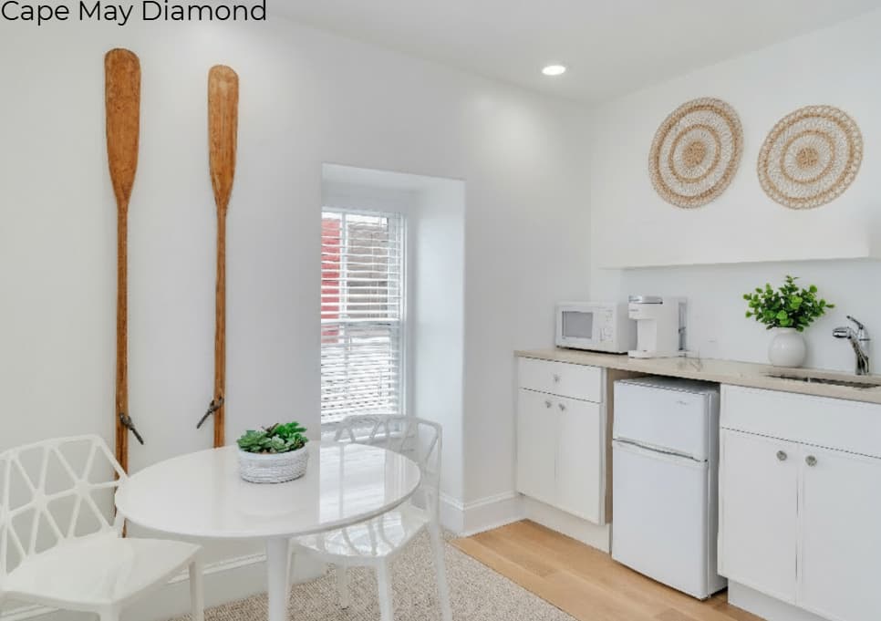 A white kitchenette area in a bright room featuring white cabinets, a light wood countertop, a small refrigerator, and a microwave. To the left, a small round white dining table with two modern chairs sits beneath two decorative wooden oars mounted vertically on the wall.