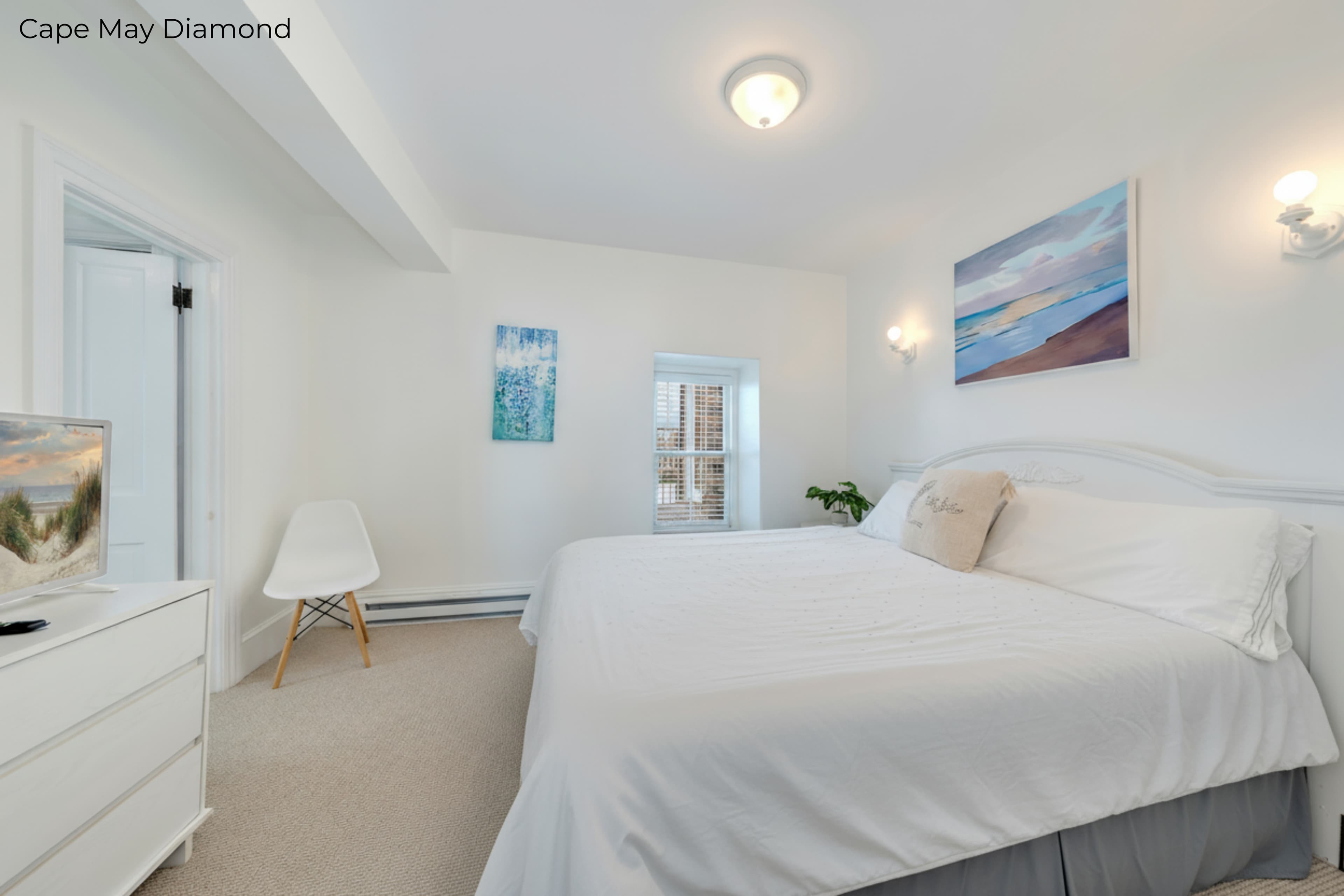A bright bedroom with white walls and beige carpeting features a bed dressed in crisp white linens and a grey bedskirt. Above the arched white headboard hangs a horizontal painting of a beach sunset, flanked by two glowing wall sconces. To the left, a white dresser and a modern white chair are visible near an open doorway.