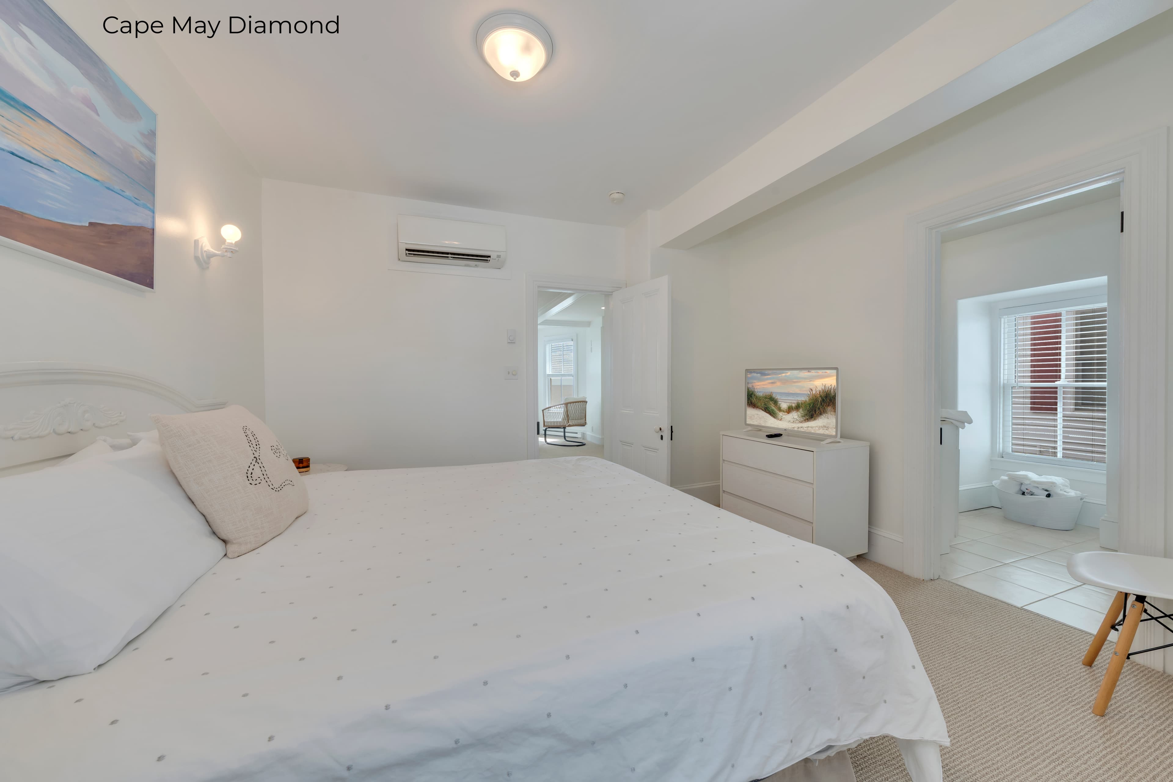 A bright, modern bedroom featuring a bed with crisp white linens and a grey bedskirt. A coastal sunset painting hangs above the headboard, flanked by two illuminated wall sconces. To the left, a white dresser holds a small television, and an open doorway leads to another bright room with a window.