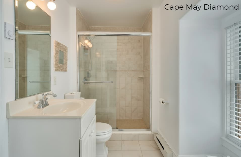 A brightly lit bathroom with white walls and light tan floor tiles. The space features a white vanity with a chrome faucet and a large mirror, a white toilet, and a glass-enclosed walk-in shower with beige tiles. To the right, a window with white horizontal blinds provides natural light.