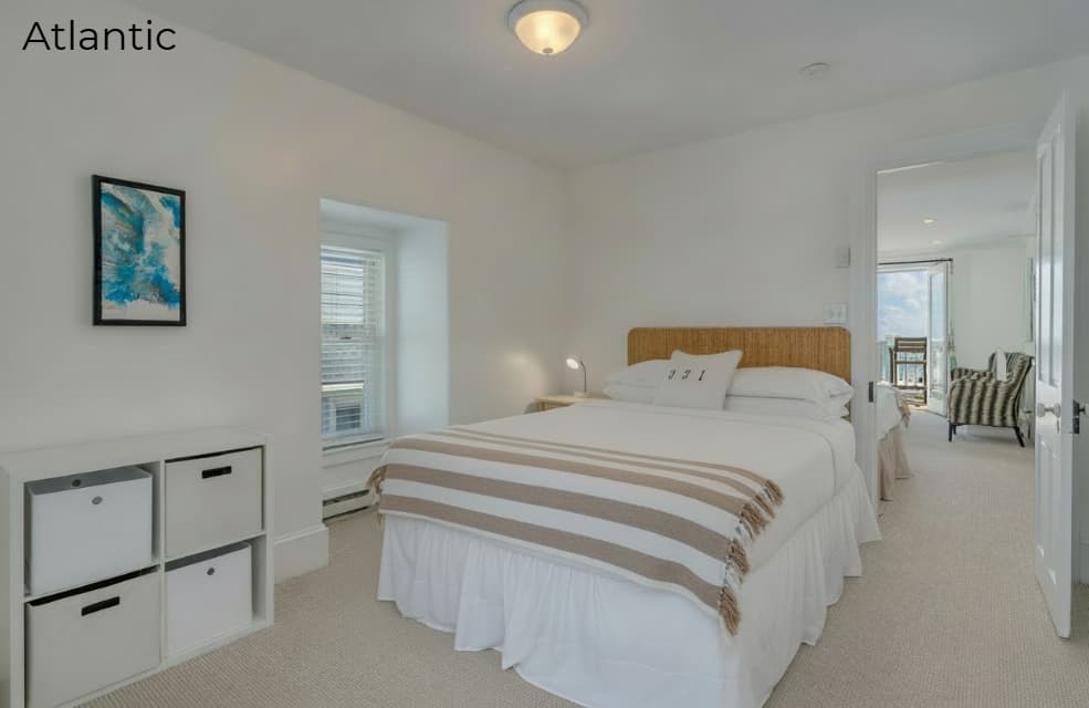 A brightly lit, modern bedroom featuring a bed with a wooden headboard, white linens, and a striped throw blanket. To the left, a white cube storage unit sits against the wall beneath a blue abstract painting, while an open doorway on the right reveals a sunlit living area with a view of the ocean.