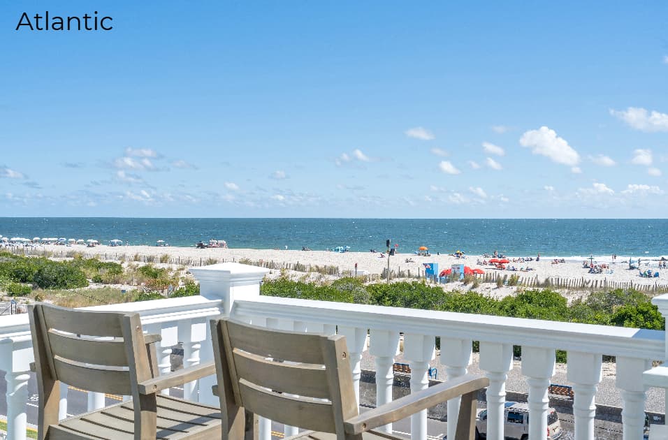 A wide-angle view from a balcony with two wooden chairs overlooking a bright, sunny beach. Beyond the white railing, the scene features sandy dunes with green shrubs, a populated beach with colorful umbrellas, and a calm blue ocean under a clear sky with scattered white clouds.