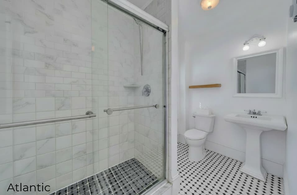 A bright, modern bathroom featuring black and white patterned floor tiles and white walls. On the left, a large walk-in shower is enclosed by sliding glass doors and lined with white marble-style tiles. The right side of the room includes a white pedestal sink with a framed mirror above it, a white toilet, and a simple wooden floating shelf.